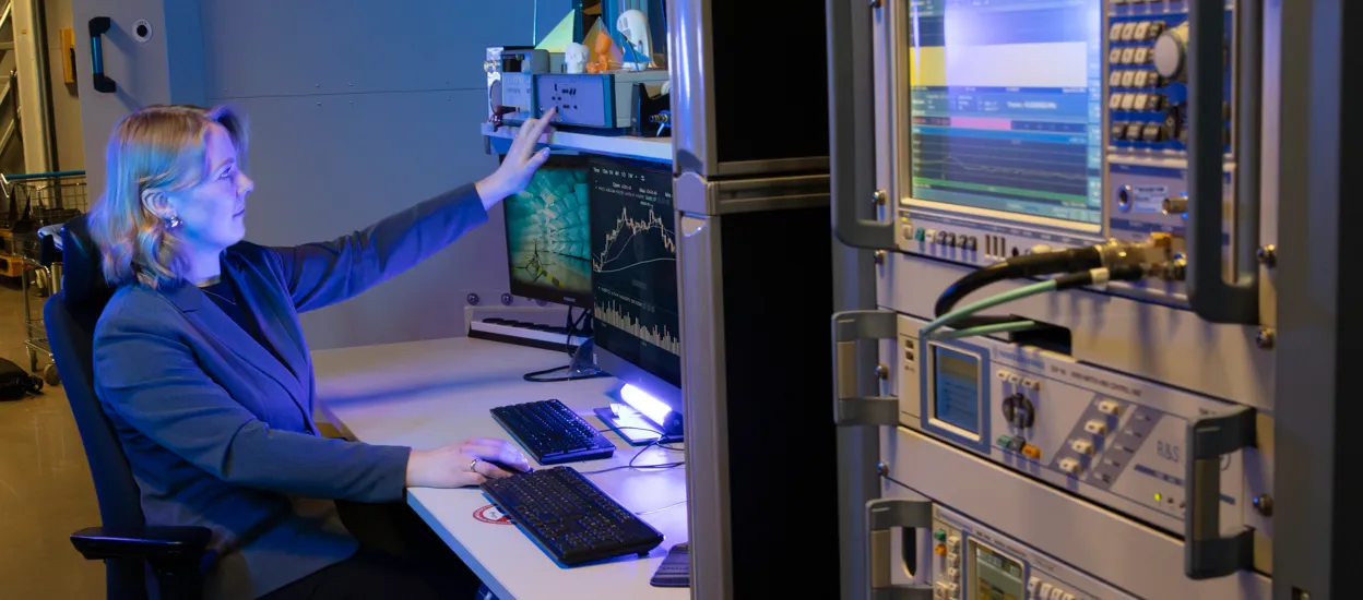 A woman is sitting at a desk in an electronics lab, with a computer screen in front of her. One hand is placed on a mouse and the other one is pressing a button on a machine above the screen. Photo.